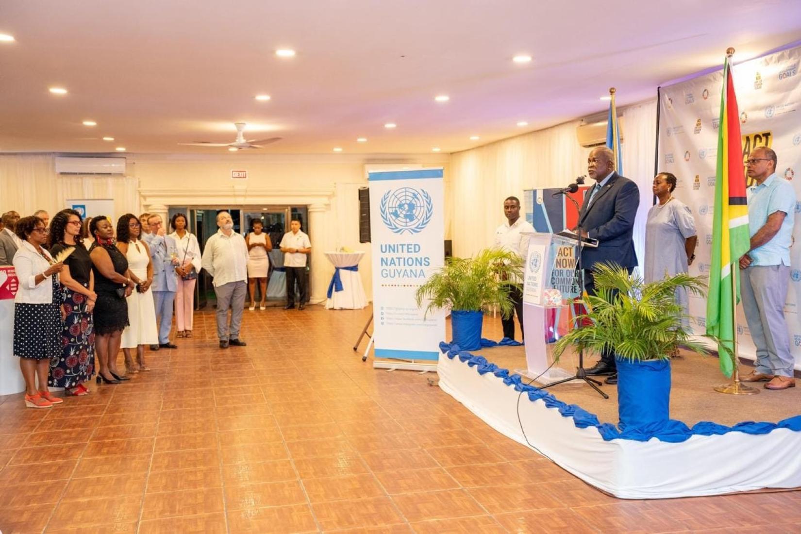 A man standing at a podium speaking with a woman and man behind him. A crowd of people are listenign to his speech.