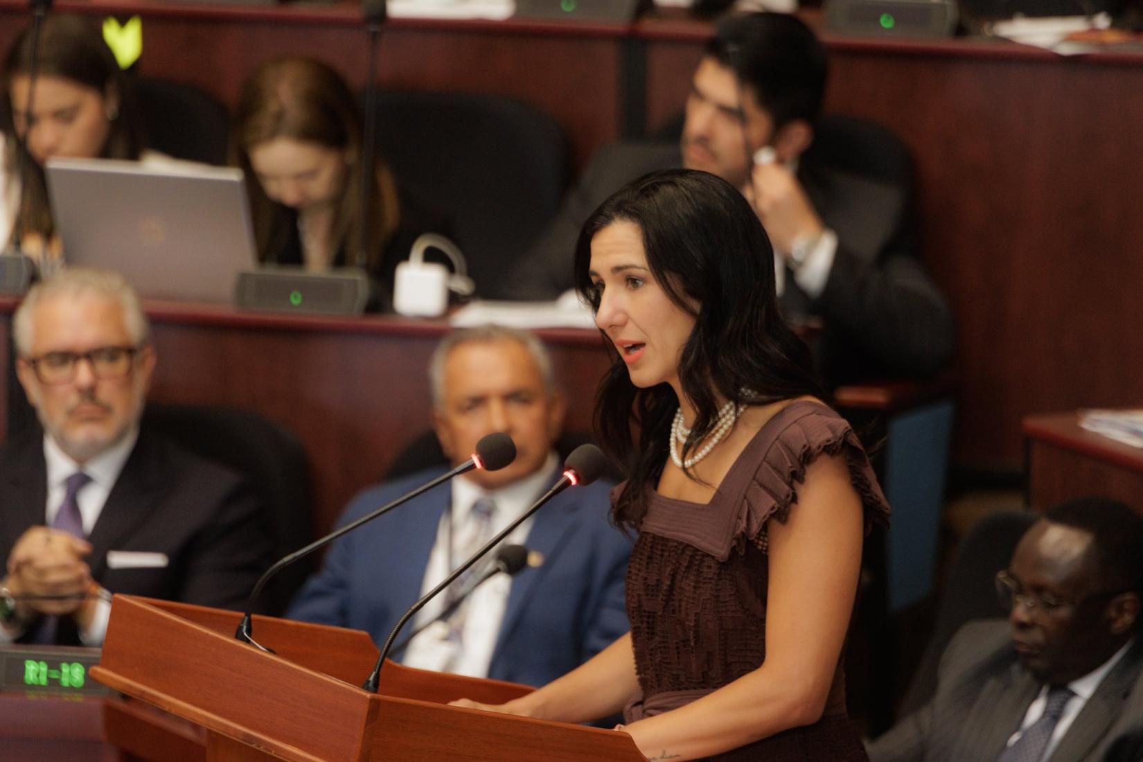 A woman standing at a podium speaking with persons seated in the background.