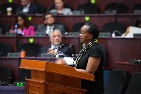 A woman standing at a podium speaking with persons seated in the background.
