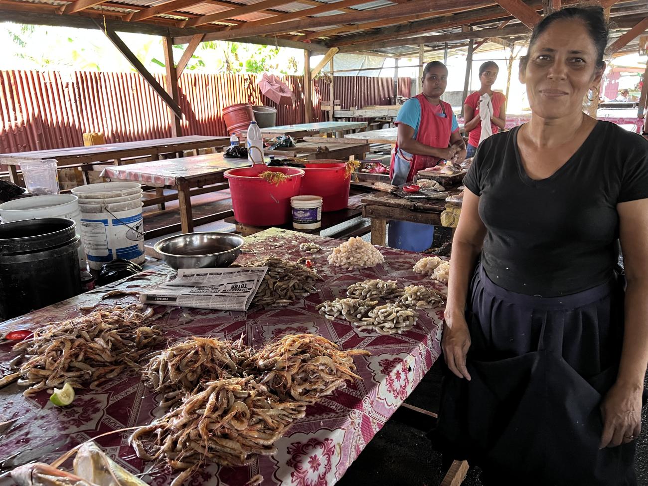 A woman standing in the foreground next to a table of shrimp. Two women standing in teh background at another table.