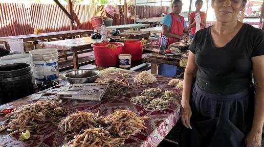 A woman standing in the foreground next to a table of shrimp. Two women standing in teh background at another table.