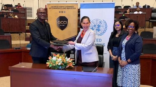 Two persons standing shaking hands while holding a document. Two other persons are standing observing.