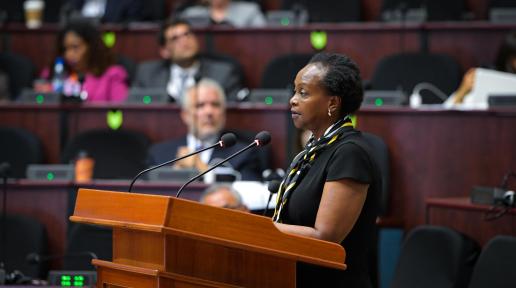 A woman standing at a podium speaking with persons seated in the background.